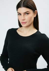 Pull noir en tissu texturé, avec un col rond et des manches longues. La coupe est ajustée, mettant en avant un design simple.