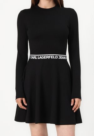 Žena v černých dlouhých rukávech a bílé pásce v pase s nápisem "KARL LAGERFELD JEANS".