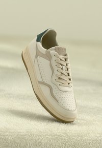 Sneaker de couro bege com biqueira perfurada, detalhes em camurça, atacadores creme, tab superior em verde escuro e sola de borracha sobre uma superfície texturizada bege.