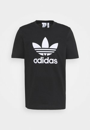 Sort T-shirt med korte ærmer med stort hvidt Adidas trefoil-logo og "adidas" tekst centreret på forsiden mod en hvid baggrund.