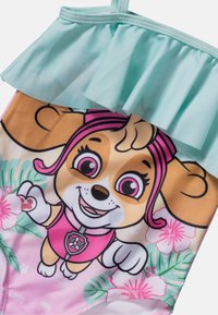 Maillot de bain une pièce avec un design de chien de dessin animé dans des couleurs rose et beige, des volants vert menthe, et des motifs de fleurs tropicales.