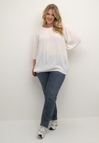 Hvid, tekstureret bluse med korte, puffede ærmer, parret med blå jeans og mønstrede sko. Let, luftig stof med diskrete detaljer.