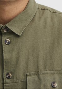 Camicia verde oliva con colletto a bottoni, caratterizzata da grandi bottoni marroni e taschino sul petto. Realizzata in un tessuto morbido e testurizzato.