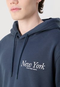 Mørkeblå hettegenser med frontlomme, har snorer og brodert "New York Calvin Klein Jeans" tekst på brystet. Myk tekstur.