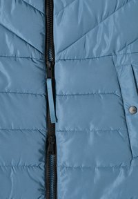 Veste matelassée bleu clair avec un design en biais, dotée d'une fermeture éclair noire, d'un tirage bleu et d'une poche latérale avec un bouton-pression.