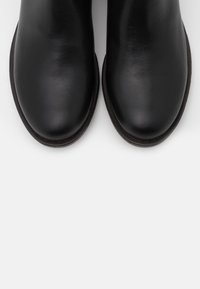 Botas de tobillo de cuero negro con textura suave, diseño de punta redondeada y detalles cosidos en la suela, con una silueta de perfil bajo.