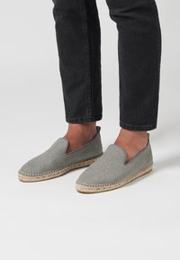 Zapatos de lona grises sin cordones con un upper texturizado, que cuentan con una suela de yute beige, punta redonda y sin cierres visibles.