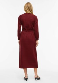 Bordowa sukienka z długim rękawem, dopasowana w talii, wykonana z fakturalnej tkaniny o prostym kroju. Zawiera dopasowany pasek do regulacji.