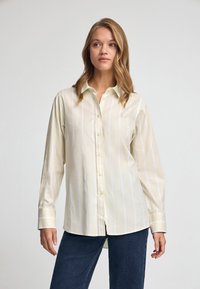 Camicia button-up gialla chiara con righe verticali bianche, maniche lunghe e un piccolo logo ricamato sul petto. Tessuto in cotone, vestibilità rilassata.