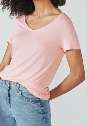 Mujer con camiseta de cuello en V color rosa claro metida en jeans de mezclilla azul de cintura alta, con la mano apoyada en la cadera.