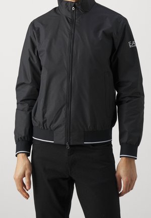 Mężczyzna noszący czarną bomberkę zapinaną na zamek z elastycznymi mankietami i dołem, białymi detalami oraz logo EA7 na lewym rękawie.
