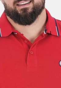Polo rouge en tissu texturé avec un col à rayures bleues et blanches, deux boutons et un patch logo sur la poitrine.