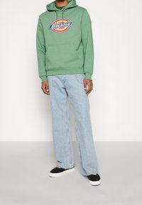 Sudadera de algodón verde con un logo central, combinada con jeans de pierna ancha de color azul claro y zapatillas negras con suelas blancas.
