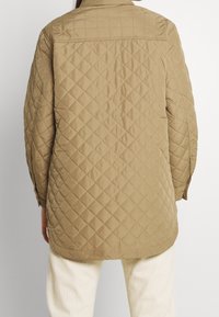 Steppjacke aus hellbraunem Stoff mit Diamantmuster, langen Ärmeln und einem Kragen. Gestaltet mit geradem Schnitt und Knopfdetails.