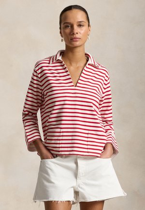Femme portant un haut à manches longues rayé rouge et blanc avec un col et un short en denim blanc aux bords effilochés, mains dans les poches.