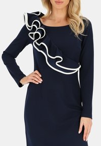 Granatowa sukienka z długimi rękawami, z falbaniastym przodem ozdobionym białym wykończeniem. Gładka tekstura, dopasowana sylwetka.