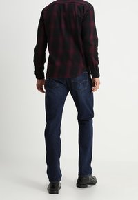 Camisa a cuadros en negro y burdeos, combinada con jeans de mezclilla azul. Los jeans tienen un corte recto y costuras sutiles en los bolsillos traseros.