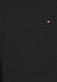Schwarzer Stoff mit glatter Textur, ausgestattet mit einer einzigen, gerade genähten Gesäßtasche und einem kleinen rot-weiß-blauen Logoakzent.