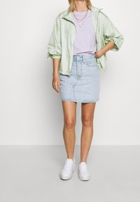 Giacca verde chiaro, sgualcita, con zip sopra una maglietta lavanda, abbinata a una gonna di jeans azzurro chiaro e scarpe da ginnastica bianche.