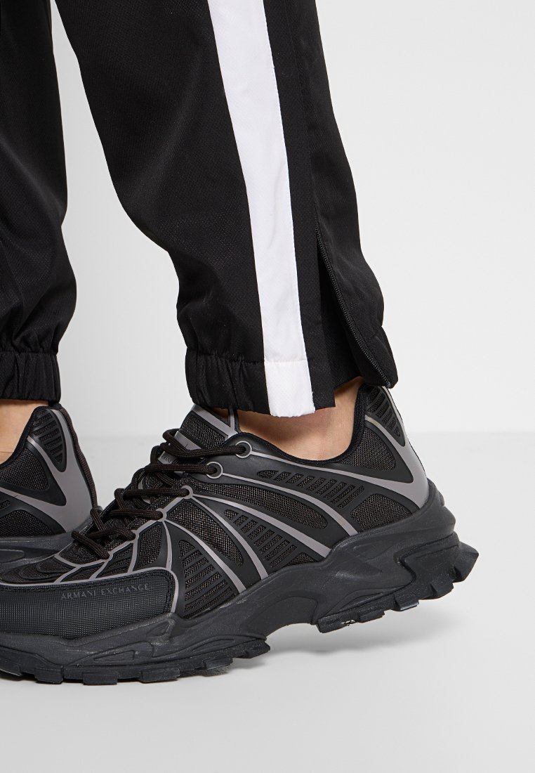 Chaussures de sport noires avec une texture en maille et en caoutchouc, présentant un design unique, des accents gris et des semelles robustes, associées à un pantalon de jogging noir.