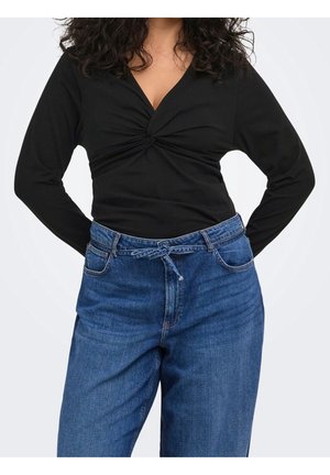 Vrouw draagt een zwarte top met lange mouwen en gedraaide voorkant en blauwe high-waist jeans met een gestrikte riem, handen rusten op haar heupen.