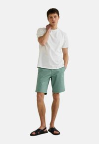 T-shirt en coton blanc, shorts vert menthe avec poches, et sandales noires. Le mannequin se tient avec une main sur le cou et l'autre dans la poche.