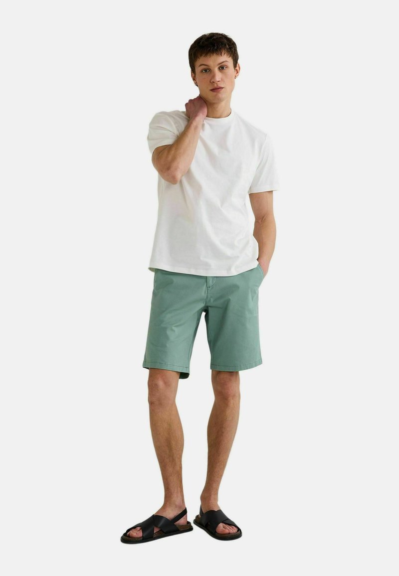 T-shirt en coton blanc, shorts vert menthe avec poches, et sandales noires. Le mannequin se tient avec une main sur le cou et l'autre dans la poche.