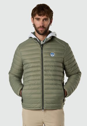 Uomo che indossa una giacca trapuntata verde chiaro con cerniera e logo circolare blu, pantaloni beige, in piedi con le mani nelle tasche su sfondo grigio.