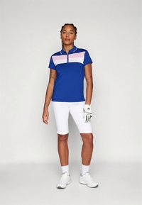 Mėlynas trumpomis rankovėmis polo marškinėliai su rožinėmis ir baltomis horizontaliomis juostelėmis, derinami su baltais šortais. Modelis dėvi baltus batus ir golfo pirštinę.