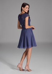Robe bleu marine avec un corsage en dentelle pailletée et une jupe en chiffon évasée, associée à des sandales à talons roses clair. Texture lisse, longueur genoux.