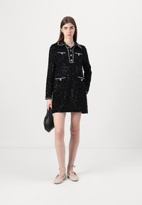 Vestido preto de lantejoulas com colarinho, dois bolsos frontais e contornos brancos. Modelo segura uma pequena bolsa preta, usando sapatos baixos bege com tiras ao tornozelo.