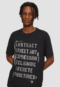 Czarny bawełniany T-shirt z dużym białym tekstem w stylu szablonu. Krótkie rękawy, okrągły dekolt, luźny krój. Tekst zawiera różne artystyczne frazy.