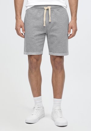 Homme portant un short gris à cordon, des chaussettes blanches et des baskets blanches, debout devant un fond clair uni.