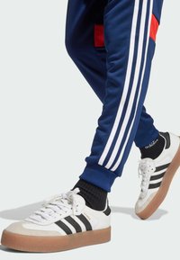 Weiße Sportsneaker mit schwarzen Streifen, Gummisohle und weißen Schnürsenkeln. Getragen mit blauen Hosen, die drei weiße Streifen und rote Akzente aufweisen.