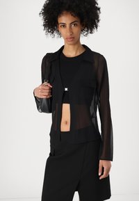 Chemisier noir transparent à manches longues avec un col, doté de deux poches poitrine et d'une coupe courte, associé à un top noir et un short.