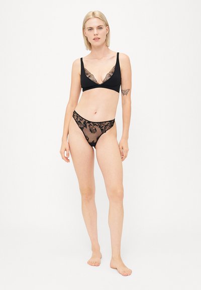 Must mustri komplekt, mis sisaldab pitsilise äärega bralette'i ning sobivat õhulist stringide komplekti lillemustriga. Mudel seisab paljajalu lihtsal taustal.