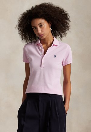 Donna con capelli ricci che indossa una polo rosa chiaro e pantaloni neri, in piedi con le mani in tasca su uno sfondo neutro.