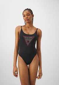 Maillot de bain une pièce noir avec un dos décolleté, présentant un design de logo triangulaire rouge et argenté avec un motif de palmier au centre.