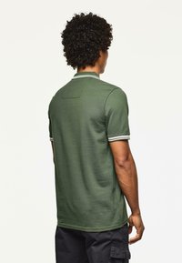 Uomo dai capelli ricci che indossa una polo a maniche corte verde scuro con colletto a righe bianche e polsini, girato di spalle.