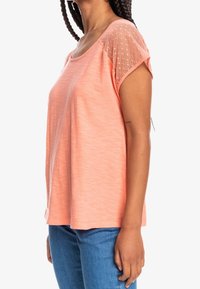 Top de color melocotón de manga corta con textura suave, con mangas de malla adornadas con un sutil patrón de puntos, corte relajado y dobladillo curvado.