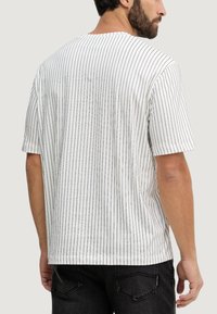 Chemise blanche à manches courtes avec des rayures verticales noires. Coupe décontractée avec un col rond et un tissu doux. Un pantalon noir est partiellement visible.