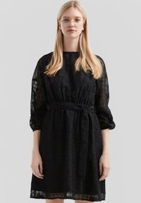 Vestido negro con un patrón floral texturizado, cintura ceñida y mangas largas de gasa. Presenta un cinturón y un corte holgado. Escote claro.