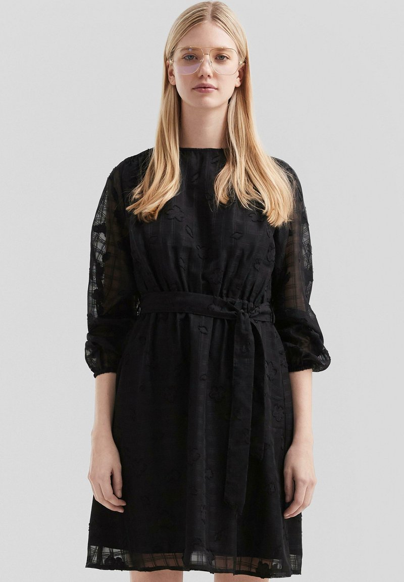 Vestido negro con un patrón floral texturizado, cintura ceñida y mangas largas de gasa. Presenta un cinturón y un corte holgado. Escote claro.