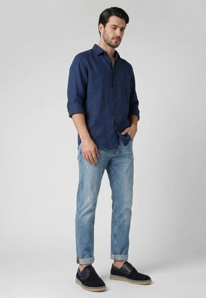 Mann trägt ein dunkelblaues Hemd mit Knöpfen, hellblaue hochgekrempelte Jeans und dunkelblaue Freizeitschuhe und steht vor einem grauen Hintergrund.