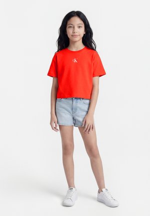 Ragazza con capelli lunghi e scuri, indossa una t-shirt rossa Calvin Klein, shorts di jeans chiari e sneakers bianche, in piedi contro uno sfondo bianco.