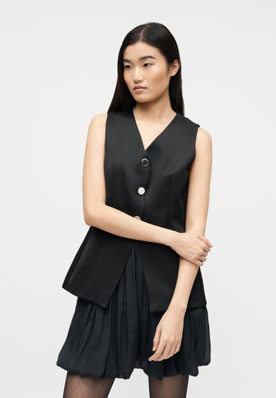 Νεαρή γυναίκα με μακριά σκούρα μαλλιά φοράει αμάνικο μαύρο γιλέκο με κουμπιά και μαύρη πλισέ φούστα, ποζάροντας με σταυρωμένα χέρια μπροστά σε απλό φόντο.