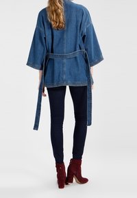 Chaqueta de kimono de denim con mangas amplias, ceñida a la cintura, que presenta un color azul medio y costuras en contraste. Se lleva con mallas oscuras y botines rojos.