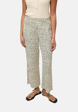 Persona que lleva pantalones crema de tiro alto y pernera ancha con puntos geométricos negros, combinados con una camiseta sin mangas beige y sandalias beige.