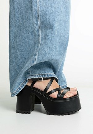 Pied portant des sandales compensées noires avec des brides croisées et une boucle, associées à un jean droit bleu clair.
