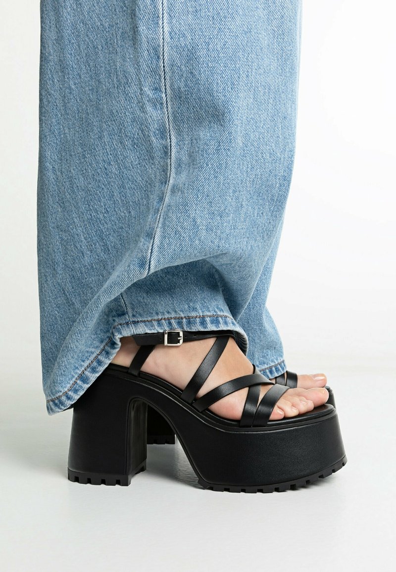 Pied portant des sandales compensées noires avec des brides croisées et une boucle, associées à un jean droit bleu clair.